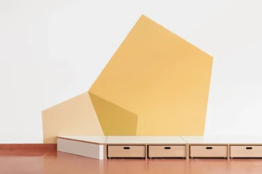 Spielpodest mit gelber Wandgestaltung Kindergartenmöbel und Raumgestaltung: Modulares Spielpodest von Papoq mit rollbaren Kisten für die einfache Aufbewahrung von Spielzeug, fördert Ordnung und Aufräumen in der Kita.