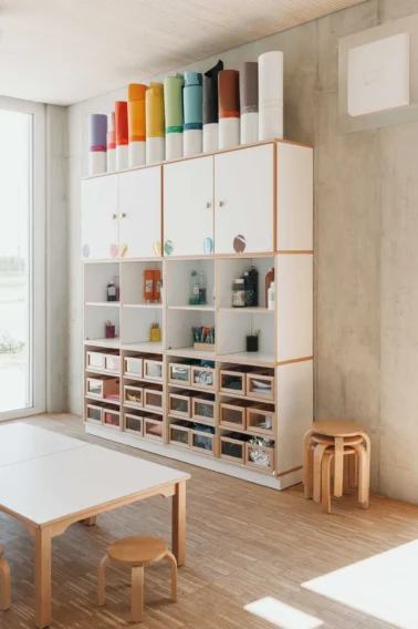 Atelier Lernwerkstatt im Kindergarten Kindergartenmöbel und Raumgestaltung: Atelier und Lernwerkstatt im Kindergarten mit farblich sortierten Materialien im Schrank, im Vordergrund ein Tisch mit Hockern für kreatives Arbeiten und Basteln.