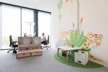 Eltern-Kind-Büro mit Spielecke Frau arbeitet am Schreibtisch im Eltern-Kind-Büro mit Kindermalecke – Papoq Möbel fördern die Vereinbarkeit von Familie und Beruf durch funktionale und durchdachte Einrichtungslösungen.